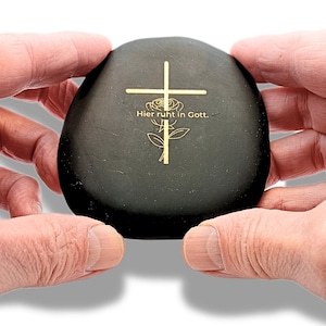 Puede incluir: Una piedra negra, lisa y redondeada, con un diseño grabado en color dorado que muestra una cruz sobre una rosa. El texto "Hier ruht in Gott." está impreso en letras doradas en el centro del gráfico de la cruz y la rosa en la piedra.