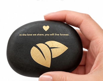 May include: A smooth, oval black stone featuring a gold-colored graphic of two leaves and a small heart. The text "In the love we share, you will live forever." is printed in gold. Above the stone, the text "PSALM Spiritual Stones" is visible.