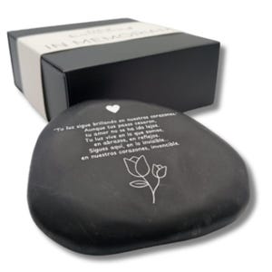 Op de afbeelding: Een gladde zwarte steen met een wit hartsymbool, een lijntekening van twee tulpen en de Spaanse tekst: Tu luz sigue brillando en nuestros corazones. Aunque tus pasos cesaron, tu amor no se ha ido lejos. Tu luz vive en lo que somos, en abrazos.