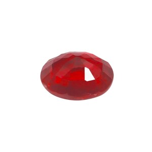May include: A faceted, oval-shaped, deep red gemstone. The gem has a polished surface with multiple facets that reflect light, creating a sparkling effect. The gemstone is set against a plain white background.