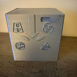Könnte beinhalten: Eine silberfarbene, quadratische Box mit geprägten Symbolen, die die Elemente Luft, Wasser, Erde und Feuer darstellen. Die Box hat auch einen Pfeil und andere geometrische Designs. Die Box ist ein dekorativer Gegenstand.