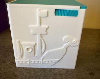 May include: White box with a teal lid and a teal accent. The box features a raised relief design of a ship. The box is on a wooden surface.