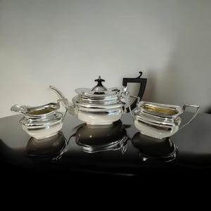 Puede incluir: Un juego de té plateado con tetera, lechera y azucarero. La tetera tiene una tapa abovedada y un pico curvo. La lechera y el azucarero tienen un diseño similar con interior dorado. El juego se exhibe sobre una superficie oscura.