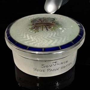 May include: A silver trinket box with a light blue and white patterned lid. The lid has a blue border and a floral design. The text "Souvenir Hyde Park Hotel" is engraved on the side.