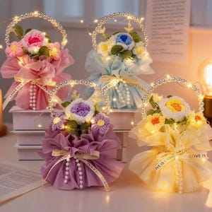 May include: Four decorative floral arrangements with LED lights. Each bouquet features crocheted flowers in pink, blue, purple, and yellow, wrapped in tissue paper and tied with a ribbon that says "LOVE IS ETERNAL". The arrangements have pearl-beaded handles.