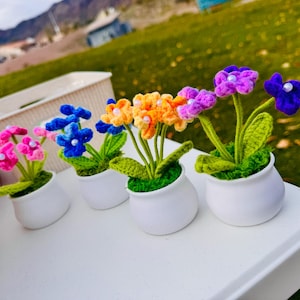 May include: A collection of handmade crochet flowers in small white pots. The flowers are in various colors, including pink, blue, orange, purple, and blue. The pots sit on a white surface with a green grassy background.