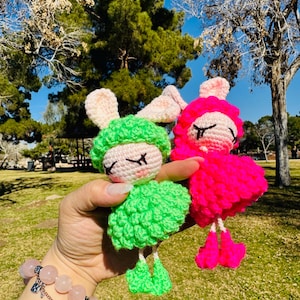 May include: Two handmade crochet bunny dolls. One doll is bright green, the other is hot pink. Both have white faces, pink cheeks, and closed eyes. The dolls have floppy ears and are holding their arms down. The background is a park with trees.