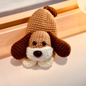May include: A handmade crochet dog toy in shades of brown, white, and black. The dog has floppy ears, a small tail, and a white muzzle. The toy is likely made of soft yarn and is designed to be a child's toy.