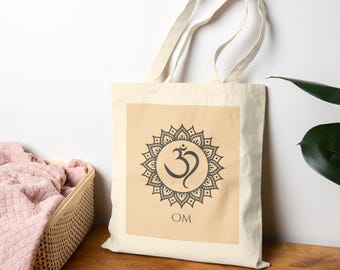 May include: A beige tote bag featuring a dark gray Om symbol within a decorative mandala design. The word "OM" is printed below the symbol. The bag has long, natural-colored handles and is made of a light-colored fabric.