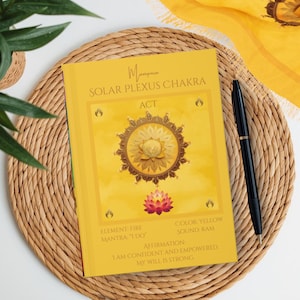 May include: A yellow book titled "Solar Plexus Chakra" with a lotus flower illustration. The book is on a woven mat with a black pen and a yellow scarf. The text on the book includes "Element Fire", "Color Yellow", and "I am confident and empowered."