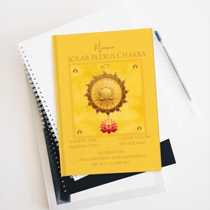 May include: A yellow journal with the words "Manipura SOLAR PLEXUS CHAKRA ACT" printed on the cover. The cover also features a gold mandala design and a pink lotus flower. The journal is on top of a white spiral notebook.