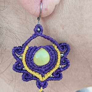 Peut inclure: Boucle d'oreille macramé faite à la main, violette et jaune. Elle présente une pierre centrale vert clair et des perles argentées. Le travail de nœuds complexe forme une forme florale, avec un fermoir argenté.
