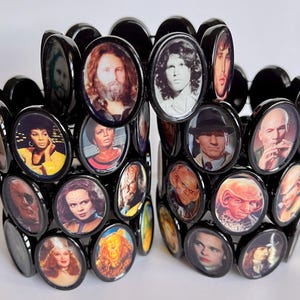 May include: A black bracelet with oval-shaped charms featuring images of various celebrities and musicians. The bracelet is made of black metal and has a unique design.