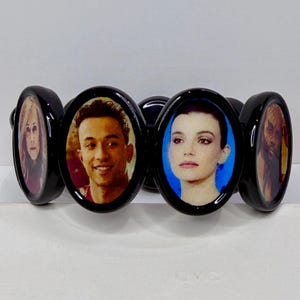 May include: A black oval-linked bracelet featuring portraits of people. Each oval has a glossy black frame. The portraits are in color and appear to be printed on the bracelet. The bracelet is on a white surface.