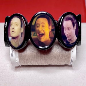 May include: A black bracelet with three round, black-rimmed charms featuring images of a man's face. The charms are arranged in a row.