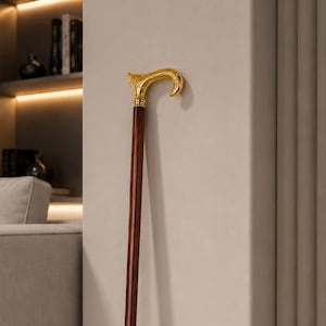 May include: A walking cane featuring a long, straight, dark brown wooden shaft and a curved, ornate gold-colored metal handle. The handle displays intricate embossed detailing, and the base of the cane is finished with a small gold-colored metal ferrule.