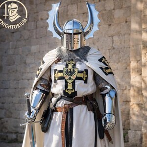 May include: A knight in full armor, including a helmet with decorative silver horns, chainmail, and a white tunic with a black cross emblem. The knight holds a long sword. The image includes the text "Knightorium" in a circular logo.
