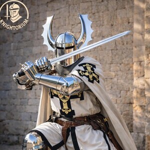 May include: A knight in full armor, holding a sword in a defensive stance. The armor is highly polished, reflecting the surroundings. The knight wears a white tunic with black trim and a cape. The helmet has decorative horns.