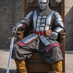 Op de afbeelding: Een persoon in volledige middeleeuwse wapenrusting, inclusief helm, mali&euml;nkolder en metalen platen, zit op een houten troon. De wapenrusting is zilverkleurig, met rode en grijze accenten. De persoon houdt een zwaard vast. Het logo "Knightorium" is zichtbaar.