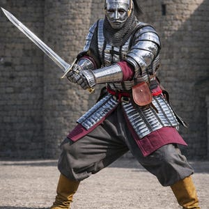 Op de afbeelding: Een persoon in volledige middeleeuwse wapenrusting, inclusief helm, mali&euml;nkolder en plaatpantser, houdt een lang zwaard vast. De wapenrusting is zilverkleurig, met bordeauxrode accenten. De persoon draagt een grijze broek en bruine laarzen.