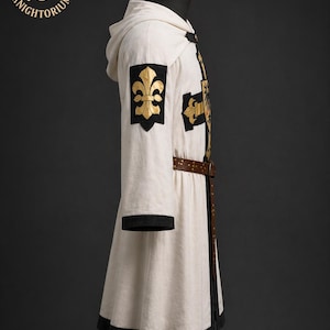 May include: A cream-colored medieval hooded robe with black trim and a brown belt. The robe features gold fleur-de-lis and cross emblems on the sleeves and chest. The "Knightorium" logo is visible in the upper left corner.