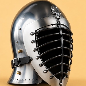 May include: A polished steel helmet featuring a black metal barred face guard, a hinged visor mechanism, and a black leather chin strap with a metal buckle. The helmet includes a logo in the upper left corner with the text KNIGHTORIUM.
