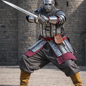 Op de afbeelding: Een persoon in volledige middeleeuwse bepantsering, inclusief helm, mali&euml;nkolder en metalen platen, houdt een lang zwaard vast. De bepantsering is zilverkleurig met bordeauxrode accenten. De persoon draagt bruine laarzen en een grijze broek. Het logo "KNIGHTORIUM" is zichtbaar.