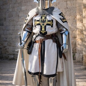 May include: A knight in full armor, including a helmet with decorative silver horns, chainmail, and a white tunic with a black cross emblem. The knight holds a long sword. The image includes the text "Knightorium".