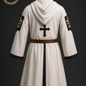 May include: A cream-colored hooded robe with black trim and a brown belt. The robe features a black cross on the back and gold fleur-de-lis patches on the sleeves. The logo "KNIGHTORIUM" is visible in the upper left corner.