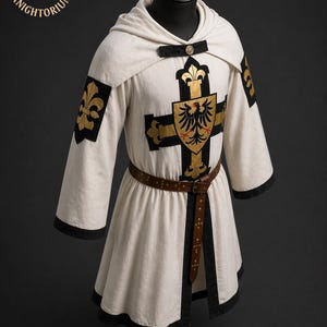 May include: Cream-colored medieval tunic with a hood, black trim, and a brown leather belt. The tunic features a black cross with a gold emblem and fleur-de-lis accents. The sleeves have gold fleur-de-lis patches. The logo "Knightorium" is in the upper left corner.