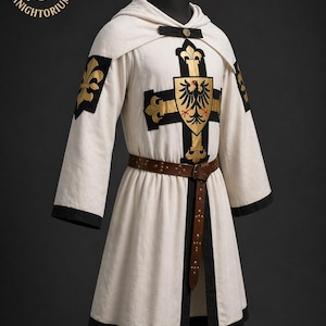 May include: A white medieval tunic with black trim, a hood, and a brown leather belt. The tunic features a gold cross design with a black eagle emblem and gold fleur-de-lis accents. The Knightorium logo is visible in the upper left corner.
