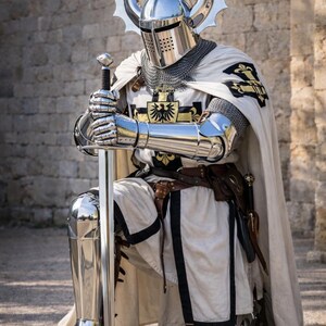 May include: A knight in full armor, kneeling and holding a sword. The armor is silver and reflective, with a helmet featuring large, decorative horns. The knight wears a white tunic and cape with black trim and heraldic emblems.