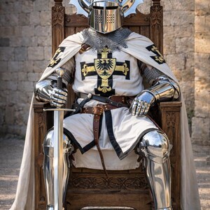 May include: A knight in full armor sits on a wooden throne, holding a sword. The armor is silver, with a white tunic featuring a black and gold cross design. A silver helmet with decorative horns and chainmail complete the look. The logo "Knightorium" is visible.