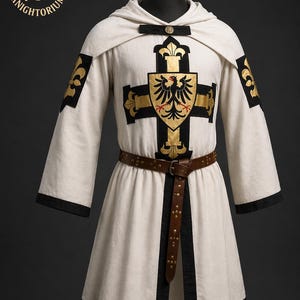 May include: Cream-colored medieval tunic with a black and gold cross design on the front. The tunic features a hood, black trim, and a brown leather belt. The sleeves have gold fleur-de-lis accents. The Knightorium logo is in the upper left corner.