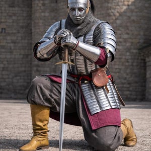 Op de afbeelding: Een persoon knielt, gekleed in gepolijst stalen harnas, mali&euml;nkolder en een helm met een gezichtsmasker. Ze houden een zwaard vast, met een leren tas aan hun zijde. De outfit omvat een bordeauxrode rok en bruine laarzen. Het beeld heeft het "Knightorium"-logo.