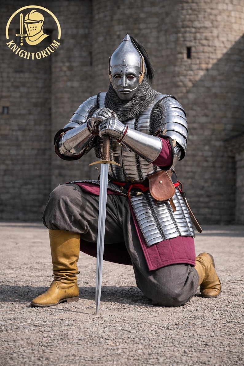 Op de afbeelding: Een persoon knielt, gekleed in gepolijst stalen harnas, mali&euml;nkolder en een helm met een gezichtsmasker. Ze houden een zwaard vast, met een leren tas aan hun zijde. De outfit omvat een bordeauxrode rok en bruine laarzen. Het beeld heeft het "Knightorium"-logo.