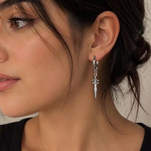 May include: A silver-toned hoop earring featuring a dangling charm shaped like a small sword or dagger. The earring is constructed from a polished metal material, with the charm suspended from a circular hoop that secures through the earlobe.