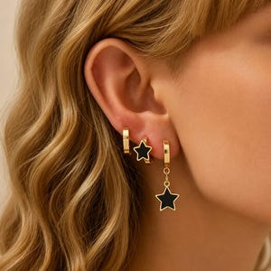 May include: Three gold-toned hoop earrings worn in a row. The first is a plain gold hoop, the second features a small black star charm attached directly to the hoop, and the third is a gold hoop with a dangling black star charm pendant.