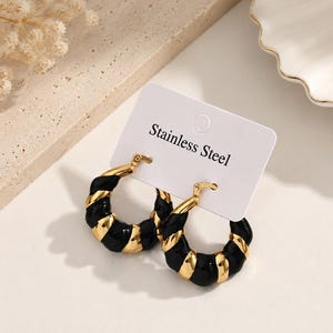 May include: A pair of hoop earrings featuring a twisted design with alternating segments of polished gold-tone metal and glossy black enamel. The earrings are attached to a white display card with the text Stainless Steel printed in black.