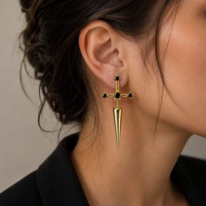 May include: A gold-toned earring shaped like a sword or dagger, featuring a cross-guard design adorned with small black stones at the center and ends, with a smooth, pointed, conical blade extending downward from the cross-guard section.