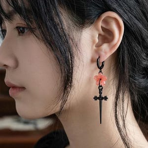 May include: A black hoop earring featuring a small, light pink five-petaled flower charm suspended above a black metal sword pendant. The earring is worn in the earlobe, showcasing a contrast between the delicate floral element and the sharp sword.