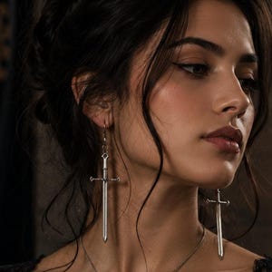 May include: A silver-toned jewelry set consisting of a necklace and a pair of earrings, each featuring a miniature sword pendant. The sword charms have a metallic finish with a cross-guard and a pointed blade design, suspended on a thin silver chain.