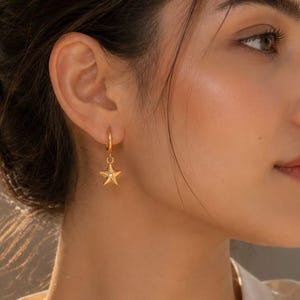 May include: A gold hoop earring featuring a small, five-pointed star charm with a tiny embedded clear stone at its center. The earring is worn in the earlobe, showcasing a polished metallic finish and a delicate, dangling design that catches light.