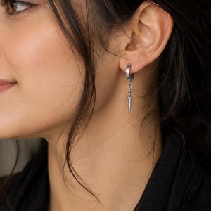 May include: A silver-toned hoop earring featuring a small, dangling dagger charm. The earring has a polished metallic finish and a secure hinged closure, with the dagger pendant hanging vertically from the bottom of the circular hoop component.