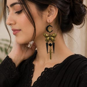 May include: A long dangle earring featuring a black crescent moon with a gold star, a gold butterfly, a black diamond-shaped charm, and a small gold sword. The earring is constructed from metallic gold and black components, hanging from a hook wire.