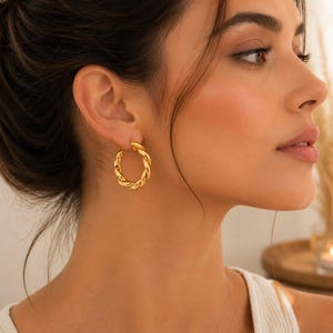 May include: A gold-toned hoop earring with a twisted, rope-like texture. The earring is circular in shape and features a polished, reflective metallic finish that catches the light, creating a distinct spiral pattern along the entire hoop surface.