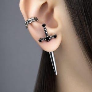 May include: A silver-toned ear cuff featuring a row of small skulls and a sword-shaped earring with black stones set into the hilt and a long, smooth, pointed blade extending downward from the earlobe.