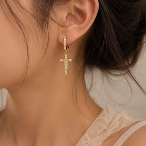 May include: A gold hoop earring featuring a dangling sword-shaped charm. The hoop and the sword charm are both encrusted with small, clear, sparkling stones. The sword charm hangs vertically from the circular hoop, which is secured in the earlobe.