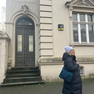 Może przedstawiać: Osoba w długim czarnym płaszczu puchowym, jasnoniebieskiej czapce i szarych spodniach dresowych. Nosi turkusową torbę na ramię z geometrycznym wzorem. W tle budynek z szarymi drzwiami i numerem 55.
