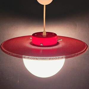 Può includere: Lampada a sospensione rossa e bianca. La lampada presenta un paralume circolare perforato e una lampadina sferica bianca. È sospesa da un cavo bianco da una base cilindrica rossa. Design retrò.
